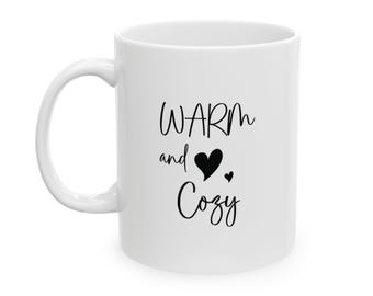 May include: White ceramic mug with a curved handle. The mug features the words "WARM and Cozy" in black script, with a black heart symbol. A small black heart is also present.