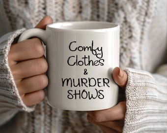 May include: White ceramic mug with the text "Comfy Clothes & Murder Shows" in black script. The mug has a curved handle and is held by a person wearing a cream-colored sweater.
