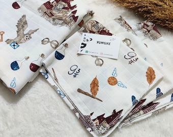 May include: Two white fabric squares with a Hogwarts theme. The fabric features a castle, owls, glasses, wands, and a golden snitch. The fabric is folded and has a red border. The fabric is on a white surface.