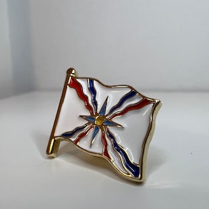Puede incluir: Un pin de color dorado con un diseño de bandera de esmalte blanco, rojo y azul. La bandera presenta una estrella estilizada con un centro amarillo.