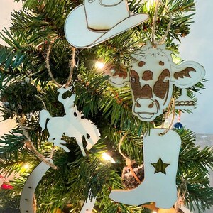 Puede incluir: Adornos navideños de madera con temática de vaquero colgados de un árbol. Los adornos incluyen un sombrero de vaquero, una cabeza de vaca, una bota de vaquero con una estrella, una herradura y una silueta de un vaquero a caballo.