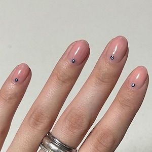 May include: Close-up of a hand with nude nail polish and small blue and white eye designs on each nail. The hand is wearing a silver ring.