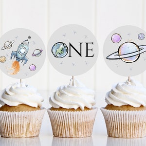 Op de afbeelding: Drie grijze en witte cupcake-toppers met een ruimtethema. De toppers tonen een raket, een planeet en het woord "ONE".