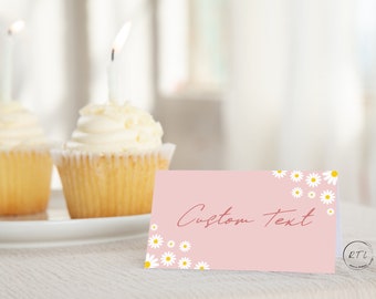 Op de afbeelding: Roze placecard met witte madeliefjes en de tekst "Custom Text" in rood gedrukt. De placecard ligt op een witte tafel met twee cupcakes op de achtergrond.