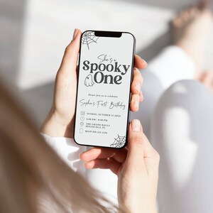 May include: A smartphone displaying a Halloween-themed first birthday party invitation. The invitation features a ghost illustration and the text "She's a Spooky One" and "Sophia's First Birthday". The invitation includes details such as the date, time, location, and RSVP information.