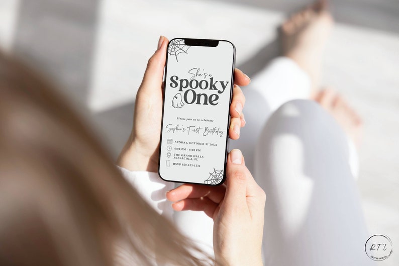 May include: A smartphone displaying a Halloween-themed first birthday party invitation. The invitation features a ghost illustration and the text "She's a Spooky One" and "Sophia's First Birthday". The invitation includes details such as the date, time, location, and RSVP information.