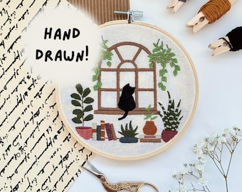 May include: An embroidery hoop featuring a hand-stitched scene of a black cat sitting on a windowsill with potted plants, books, and an arched window. The text HAND DRAWN! is visible. Includes wooden clothespins with thread and gold-toned scissors.