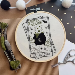 May include: An embroidery hoop featuring a tarot card design with a black cauldron, spiderweb, and the text "THE CAULDRON". The design is stitched with black and green thread on a cream-colored fabric. A string of black, orange, and white pom-poms is in the background.