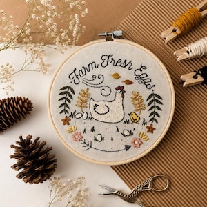 May include: A circular wooden embroidery hoop containing white fabric with hand-stitched embroidery. The design features a white hen, a yellow chick, flowers, leaves, and the text "Farm Fresh Eggs" in black thread, surrounded by pinecones and scissors.