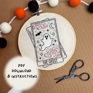 May include: An embroidered tarot card design in a wooden hoop, featuring a ghost, stars, and the text "THE GHOST". The design is in black and pink on a cream fabric. Scissors and a felt ball garland are also in the image.