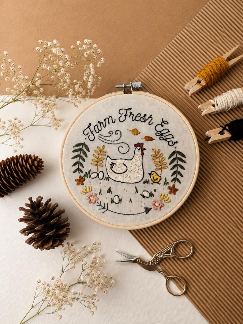 May include: A circular wooden embroidery hoop containing white fabric with hand-stitched embroidery. The design features a white hen, a yellow chick, flowers, leaves, and the text "Farm Fresh Eggs" in black thread, surrounded by pinecones and scissors.