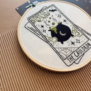 Pu&ograve; includere: Ricamo su cerchio con un design di carta dei tarocchi. La carta raffigura un calderone nero con liquido verde, un pipistrello e le parole "THE CAULDRON". Il ricamo &egrave; in un cerchio di legno, con uno sfondo di tessuto bianco. Il design include stelle e una ragnatela.
