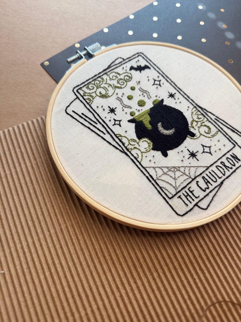 Pu&ograve; includere: Ricamo su cerchio con un design di carta dei tarocchi. La carta raffigura un calderone nero con liquido verde, un pipistrello e le parole "THE CAULDRON". Il ricamo &egrave; in un cerchio di legno, con uno sfondo di tessuto bianco. Il design include stelle e una ragnatela.