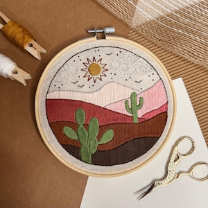 May include: A round embroidery hoop featuring a desert landscape scene. The design includes a sun, mountains, clouds, and cacti, rendered in shades of red, pink, green, and brown. The hoop is made of wood, and the scene is stitched on a cream-colored fabric.