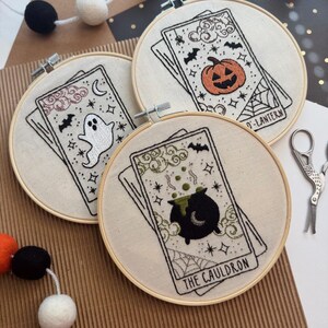 Pu&ograve; includere: Tre ricami a tema Halloween su cerchi di legno raffiguranti carte dei tarocchi. I disegni includono un fantasma, una zucca e un calderone. Il ricamo &egrave; su tessuto color crema, con accenti neri e arancioni. Sullo sfondo, una ghirlanda di palline di feltro arancioni, nere e bianche.