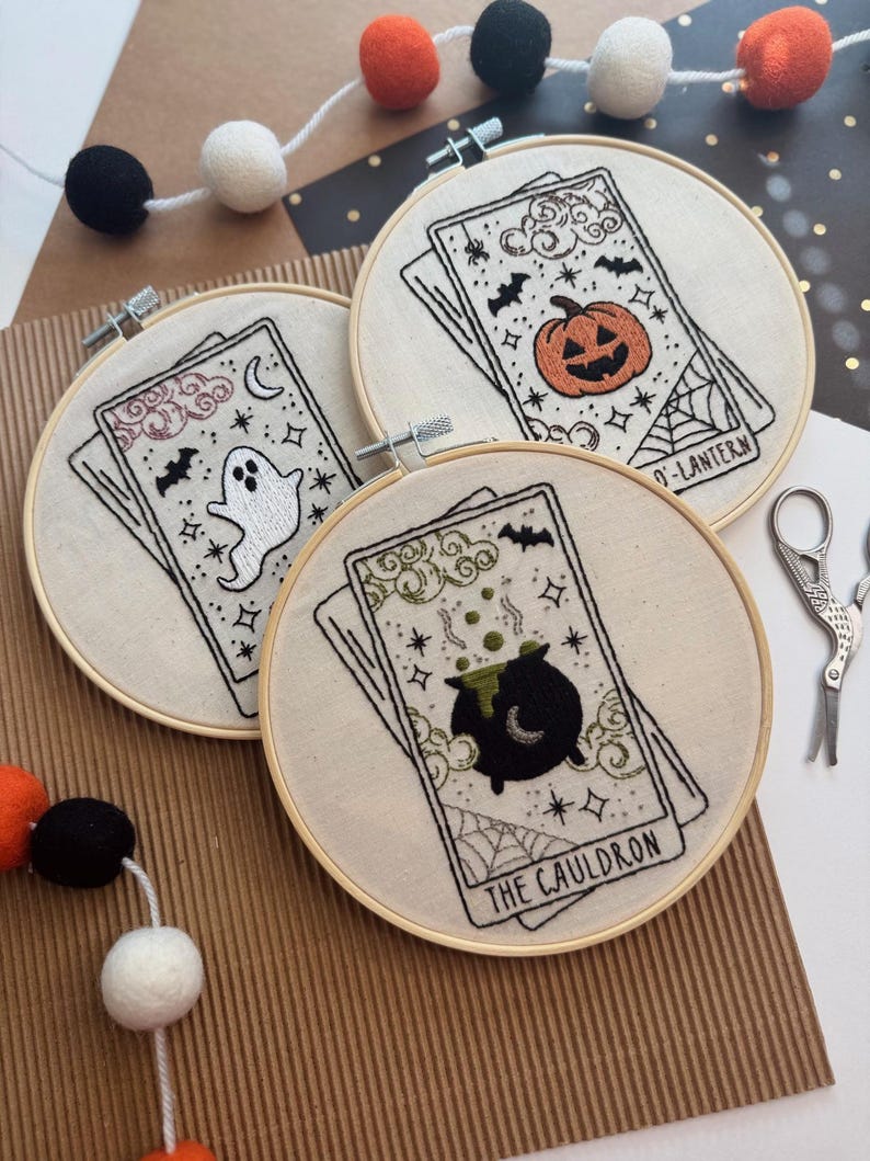 Pu&ograve; includere: Tre ricami a tema Halloween su cerchi di legno raffiguranti carte dei tarocchi. I disegni includono un fantasma, una zucca e un calderone. Il ricamo &egrave; su tessuto color crema, con accenti neri e arancioni. Sullo sfondo, una ghirlanda di palline di feltro arancioni, nere e bianche.