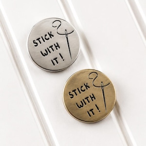 May include: Two circular metal pins, one in silver tone and one in brass tone, each featuring the engraved text "STICK WITH IT!" alongside a graphic illustration of a sewing needle with thread looping through the eye of the needle.