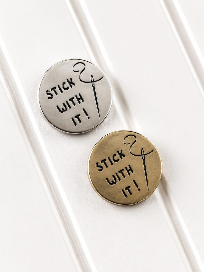 May include: Two circular metal pins, one in silver tone and one in brass tone, each featuring the engraved text "STICK WITH IT!" alongside a graphic illustration of a sewing needle with thread looping through the eye of the needle.