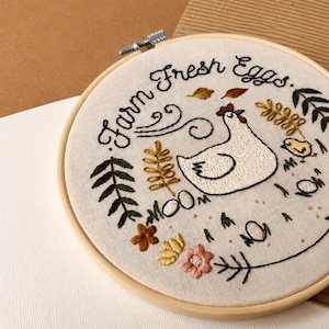 May include: A circular wooden embroidery hoop containing off-white fabric with hand-stitched embroidery. The design features a white hen, a small yellow chick, colorful flowers, leaves, and the black embroidered text "Farm Fresh Eggs."