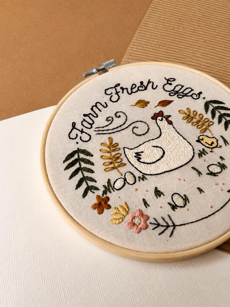 May include: A circular wooden embroidery hoop containing off-white fabric with hand-stitched embroidery. The design features a white hen, a small yellow chick, colorful flowers, leaves, and the black embroidered text "Farm Fresh Eggs."