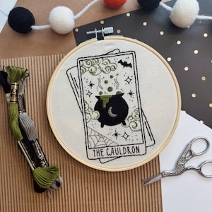 Pu&ograve; includere: Un telaio da ricamo con un disegno di carta dei tarocchi con un calderone nero, una ragnatela e la scritta "THE CAULDRON". Il disegno &egrave; ricamato con filo nero e verde su tessuto color crema. Sullo sfondo, una ghirlanda di pompon neri, arancioni e bianchi.