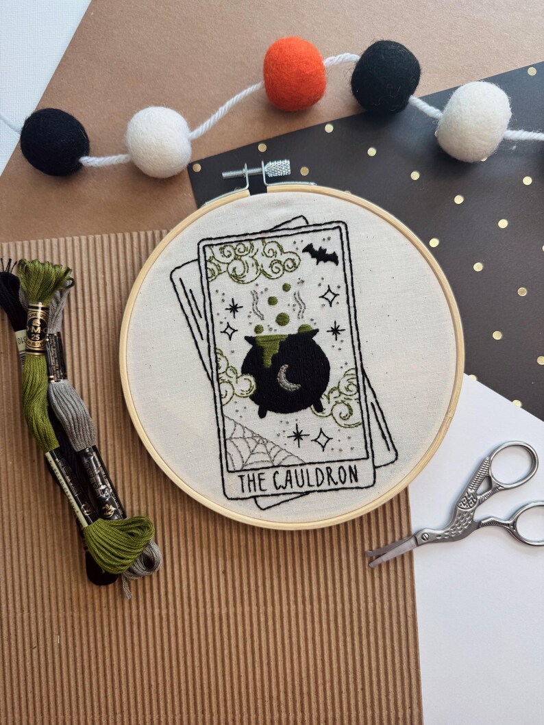 Pu&ograve; includere: Un telaio da ricamo con un disegno di carta dei tarocchi con un calderone nero, una ragnatela e la scritta "THE CAULDRON". Il disegno &egrave; ricamato con filo nero e verde su tessuto color crema. Sullo sfondo, una ghirlanda di pompon neri, arancioni e bianchi.