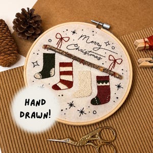 May include: A circular embroidery hoop containing fabric with a hand-stitched design of four stockings, a twig, and the text "Merry Christmas". The design includes red bows, stars, and dots. A pair of gold crane-shaped scissors sits below the hoop.