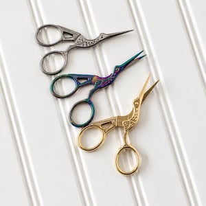 May include: Three pairs of stork-shaped embroidery scissors arranged diagonally. One pair is finished in a metallic gunmetal grey, one pair features an iridescent rainbow finish, and the third pair is finished in a polished gold tone.