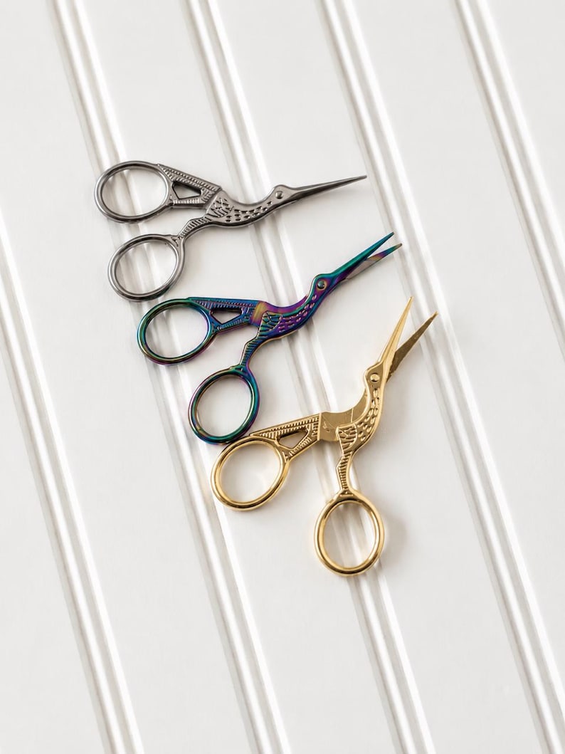 May include: Three pairs of stork-shaped embroidery scissors arranged diagonally. One pair is finished in a metallic gunmetal grey, one pair features an iridescent rainbow finish, and the third pair is finished in a polished gold tone.