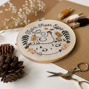 May include: A circular wooden embroidery hoop containing a fabric piece with hand-stitched designs of a hen, a chick, flowers, leaves, and the text Farm Fresh Eggs. The embroidery features black, yellow, green, and pink thread on light fabric.