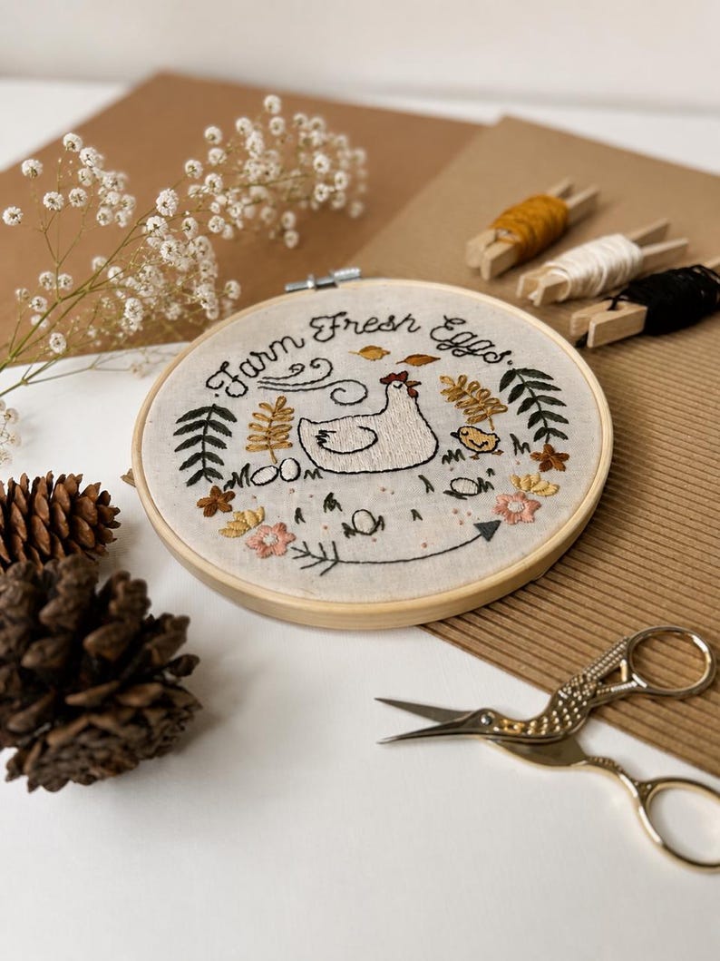 May include: A circular wooden embroidery hoop containing a fabric piece with hand-stitched designs of a hen, a chick, flowers, leaves, and the text Farm Fresh Eggs. The embroidery features black, yellow, green, and pink thread on light fabric.