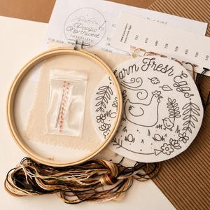 May include: An embroidery kit featuring a round wooden hoop, a piece of fabric with a printed chicken and the text Farm Fresh Eggs, a small plastic bag containing needles, a color-coded thread card, and a bundle of assorted embroidery floss.
