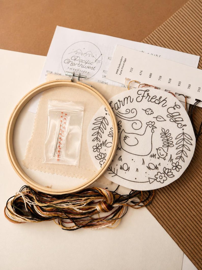 May include: An embroidery kit featuring a round wooden hoop, a piece of fabric with a printed chicken and the text Farm Fresh Eggs, a small plastic bag containing needles, a color-coded thread card, and a bundle of assorted embroidery floss.