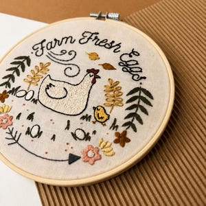May include: A circular wooden embroidery hoop containing off-white fabric with hand-stitched designs. The embroidery features a white hen, a yellow chick, floral elements, leaves, and the black text "Farm Fresh Eggs" arranged in a semi-circle.
