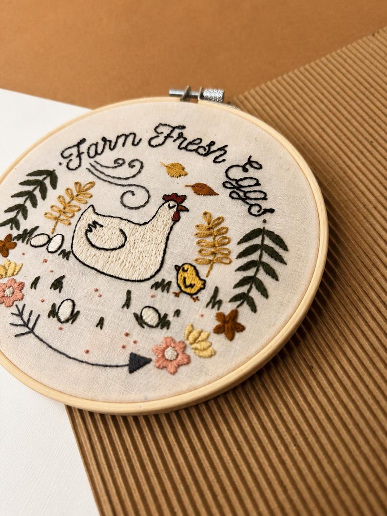 May include: A circular wooden embroidery hoop containing off-white fabric with hand-stitched designs. The embroidery features a white hen, a yellow chick, floral elements, leaves, and the black text "Farm Fresh Eggs" arranged in a semi-circle.