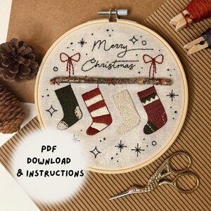 May include: A circular embroidery hoop containing a fabric piece with hand-stitched designs of four Christmas stockings hanging from a twig. The text Merry Christmas is stitched above the stockings, surrounded by small star and dot motifs.