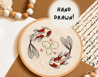 May include: A circular wooden embroidery hoop containing a fabric panel with hand-stitched koi fish in red and black thread. The design features two koi swimming in a circle around green lily pads and small pink flowers, with the text HAND DRAWN!