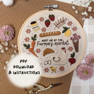 May include: Embroidered hoop art featuring a farmer's market theme. The design includes carrots, strawberries, a loaf of bread, a croissant, a hen, and the text "Meet Me At The Farmers Market." The hoop is held in a wooden frame.