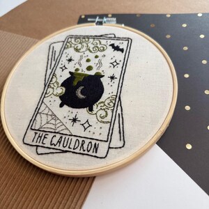 Pu&ograve; includere: Ricamo di una carta dei tarocchi con un calderone nero, una luna crescente, bolle verdi e stelle. La carta recita "THE CAULDRON". Il ricamo &egrave; posto in un cerchio di legno, con sfondo in tessuto color crema.