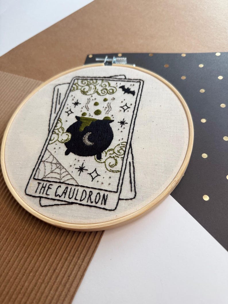 Pu&ograve; includere: Ricamo di una carta dei tarocchi con un calderone nero, una luna crescente, bolle verdi e stelle. La carta recita "THE CAULDRON". Il ricamo &egrave; posto in un cerchio di legno, con sfondo in tessuto color crema.