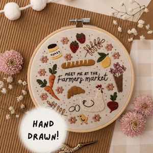 May include: A circular wooden embroidery hoop containing a beige fabric panel with hand-stitched designs of a jar of honey, strawberries, a baguette, a lemon, carrots, a croissant, a chicken, a chick, and flowers with the text MEET ME AT THE Farmers market.
