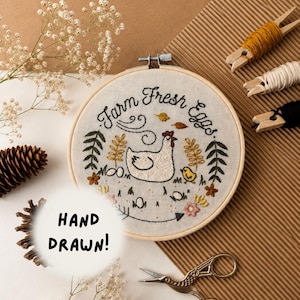 May include: A circular wooden embroidery hoop containing fabric with hand-stitched embroidery of a white hen, a yellow chick, and the text "Farm Fresh Eggs". The design includes green leaves, small flowers, and brown leaves. A white circle overlay says "HAND DRAWN!".