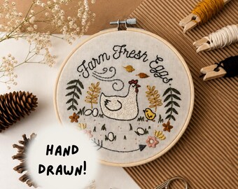 May include: A circular wooden embroidery hoop containing fabric with hand-stitched embroidery of a white hen, a yellow chick, and the text "Farm Fresh Eggs". The design includes green leaves, small flowers, and brown leaves. A white circle overlay says "HAND DRAWN!".