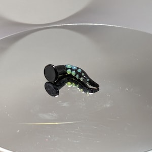 Könnte beinhalten: Ein schwarzer Glas-Ohrstöpsel mit einer geschwungenen Form und einem Design aus grün-blauen opalartigen Punkten.
