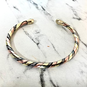 May include: A twisted metal bracelet with alternating bands of gold, silver, and rose gold. The ends of the bracelet are capped with gold-colored metal. The bracelet is displayed on a white and gray marble surface.