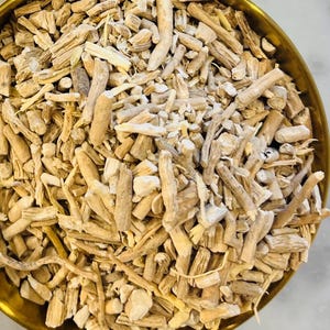 May include: A close-up of a brass bowl filled with light brown, dried, and chopped roots. The roots vary in size and shape, with some appearing like small sticks. The bowl is round and has a golden hue.