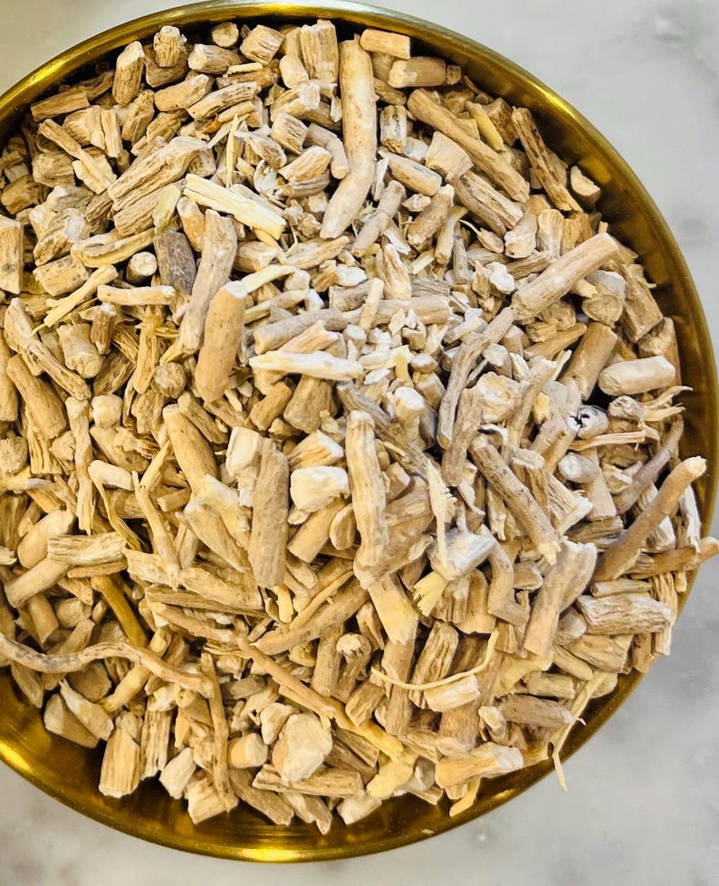 May include: A close-up of a brass bowl filled with light brown, dried, and chopped roots. The roots vary in size and shape, with some appearing like small sticks. The bowl is round and has a golden hue.