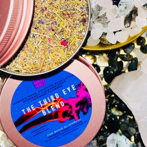 Op de afbeelding: Een roze metalen blikje met een blauw etiket met de tekst "The Third Eye Blend" met de tekst "For Mood Alignment". Het blikje bevat een mengsel van gedroogde kruiden en bloemen. De afbeelding toont ook kristallen en stenen.