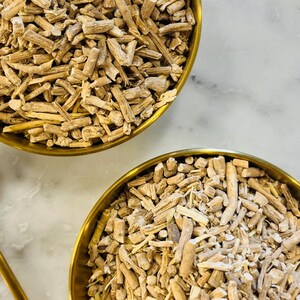 May include: Two gold-coloured bowls filled with light brown, chopped, dried roots. The roots have a fibrous texture and vary in size. The bowls sit on a white and grey marble surface. The image is a close-up shot.