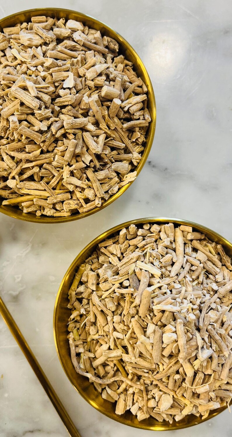 May include: Two gold-coloured bowls filled with light brown, chopped, dried roots. The roots have a fibrous texture and vary in size. The bowls sit on a white and grey marble surface. The image is a close-up shot.